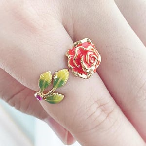 Puede incluir: Un anillo dorado con una rosa roja y hojas verdes con detalles amarillos. La rosa tiene un borde dorado y una gema rosa en la base de las hojas. El anillo es de diseño floral.