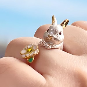 May include: Two gold rings, one featuring a white rabbit and the other a white flower with a green stem and a yellow center.
