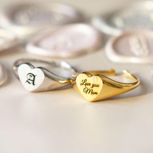 May include: Two heart-shaped signet rings. One is silver with a white heart and the letter "A". The other is gold with a heart engraved with "Love you Mum". The rings are on a white surface.