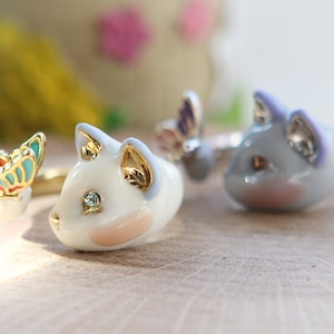May include: Two gold-tone rings featuring a white cat with a butterfly and a light purple cat with a butterfly.