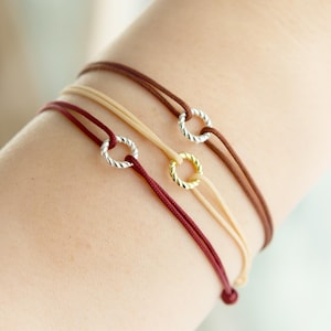 May include: Three bracelets with adjustable cords and a small twisted metal ring charm. The bracelets are in burgundy, beige, and brown.