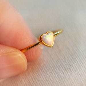 Puede incluir: Un anillo dorado con una piedra preciosa de ópalo en forma de corazón. El ópalo tiene una apariencia blanca e iridiscente, engastado en una delicada banda dorada. El anillo está sostenido por los dedos de una persona, mostrando el diseño de la joya.