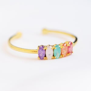 May include: A gold bangle bracelet with a row of colorful oval-shaped stones. The stones are purple, white, green, orange, and pink.