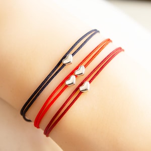May include: Three red and one black string bracelets with silver heart charms. The bracelets are layered on a person's wrist.