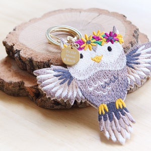 Puede incluir: Un llavero de búho bordado en gris y blanco con un anillo dorado y una etiqueta dorada que dice "To the Moon & Back". El búho lleva una corona de flores con flores rosas, amarillas y verdes.
