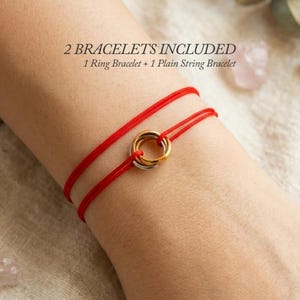 Könnte beinhalten: Zwei rote Schnurarmbänder mit einem zentralen Metallring-Anhänger. Der Text 2 BRACELETS INCLUDED 1 Ring Bracelet + 1 Plain String Bracelet ist sichtbar. Die Armbänder bestehen aus dünnen, leuchtend roten Kordeln, die um das Handgelenk liegen.