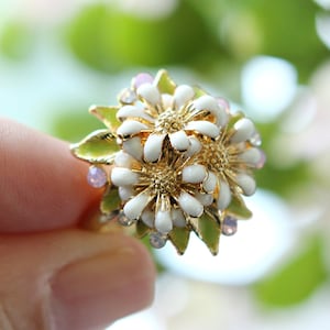 May include: A gold ring featuring a cluster of white enamel daisies with green leaves and pink rhinestones.