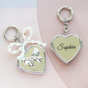 May include: Silver heart-shaped locket keychain with a pearl chain and a bow on the front. The locket has the name "Sophia" engraved on the inside.