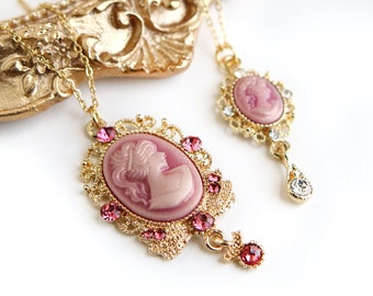 May include: Two gold necklaces with pink cameo pendants. The larger pendant features a detailed ornate frame with pink crystals. The smaller pendant has a simpler frame with clear crystals.