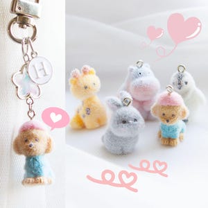 May include: A keychain with a small, plush poodle charm wearing a blue sweater and pink hat. Other plush animal charms include a giraffe, hippo, bunny, and penguin. The keychain has a flower and a circle with the letter "H".