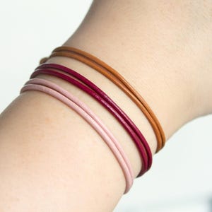 Könnte beinhalten: Drei Lederarmbänder in Rosa, Burgunderrot und Braun, die auf dem Handgelenk einer Person übereinander liegen.