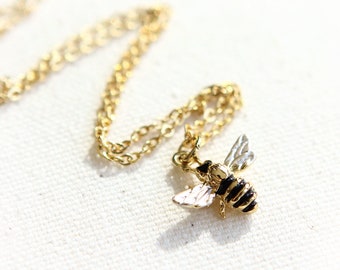 May include: A gold chain necklace with a bee pendant. The bee is black and gold with white wings.