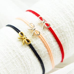 Può includere: Tre delicati braccialetti con ciondoli a forma di fiocco. Un braccialetto ha un fiocco dorato su un cordino nero, un altro ha un fiocco argentato su un cordino color pesca e il terzo ha un fiocco argentato su un cordino rosso. I braccialetti sono esposti su una superficie bianca strutturata.