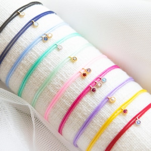 Op de afbeelding: Een verzameling kleurrijke draadarmbanden met kleine, goudkleurige bedels. De armbanden zijn er in verschillende kleuren, waaronder zwart, blauw, groen, roze, paars, geel en rood. Elke armband heeft een delicaat, minimalistisch ontwerp.