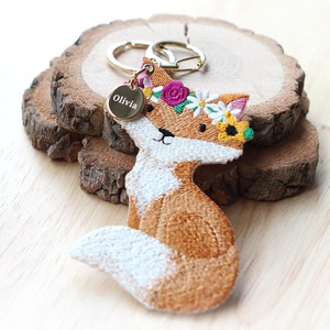 May include: A brown fox keychain with a white tail and a flower crown. The keychain has a gold tag with the name "Olivia" engraved on it.