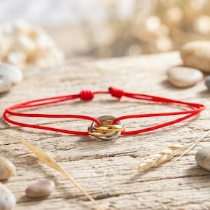 May include: A bracelet featuring a thin red cord with adjustable sliding knots and a central charm composed of three interlocking rings in silver, yellow gold, and rose gold tones, creating a layered metallic effect on the cord.