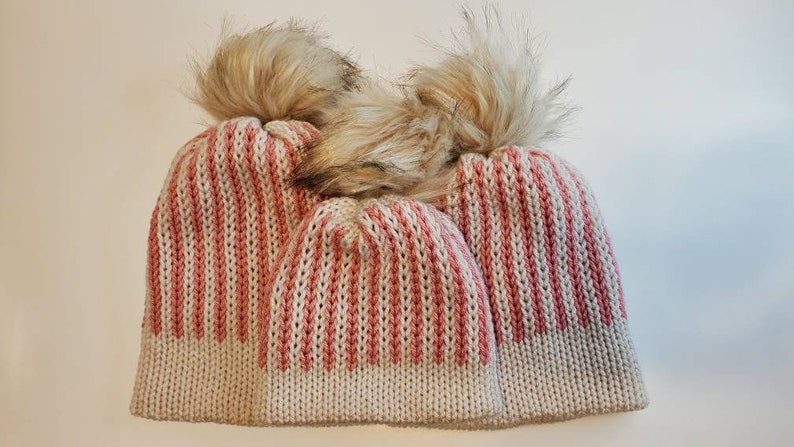 May include: Three knitted winter hats with a ribbed pattern in cream and dusty rose. Each hat is topped with a fluffy, light brown faux fur pom-pom. The hats are arranged in a group, suggesting a family set.