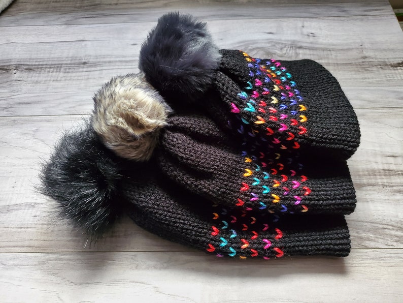 K&ouml;nnte beinhalten: Schwarze Strickm&uuml;tze mit drei bunten Bommeln. Die M&uuml;tze hat ein Regenbogen-Herz-Muster.