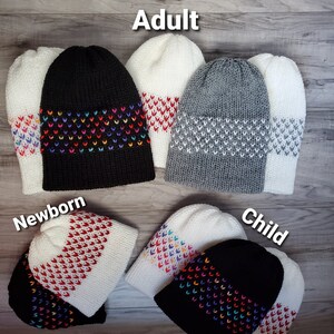 K&ouml;nnte beinhalten: Ein Set aus sechs gestrickten M&uuml;tzen in verschiedenen Farben und Gr&ouml;&szlig;en, beschriftet mit "Erwachsene", "Neugeborene" und "Kind". Die M&uuml;tzen haben ein wei&szlig;es Band mit einem bunten Herzmuster.