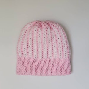 May include: A knitted beanie hat in a two-tone design. The bottom band is a solid light pink, transitioning to a striped pattern of white and light pink. The hat is made of a soft, textured yarn.