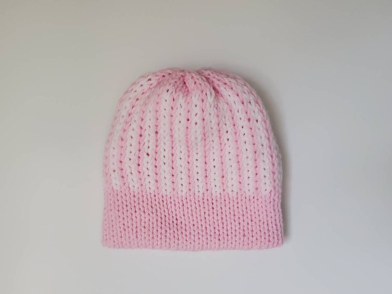 May include: A knitted beanie hat in a two-tone design. The bottom band is a solid light pink, transitioning to a striped pattern of white and light pink. The hat is made of a soft, textured yarn.