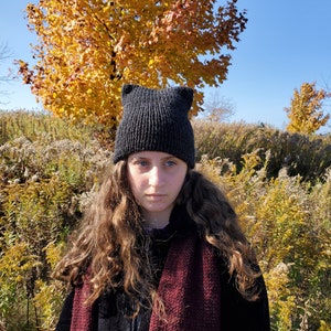 May include: A person wearing a black knit hat with cat ears and a burgundy scarf. The hat is pulled down over their forehead and the scarf is wrapped around their neck. The person is standing in front of a background of yellow and brown foliage.