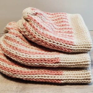 May include: A stack of three hand-knitted hats in a light beige and pink striped pattern. The hats are made of thick yarn and appear soft and warm. The hats are folded and stacked on top of each other.
