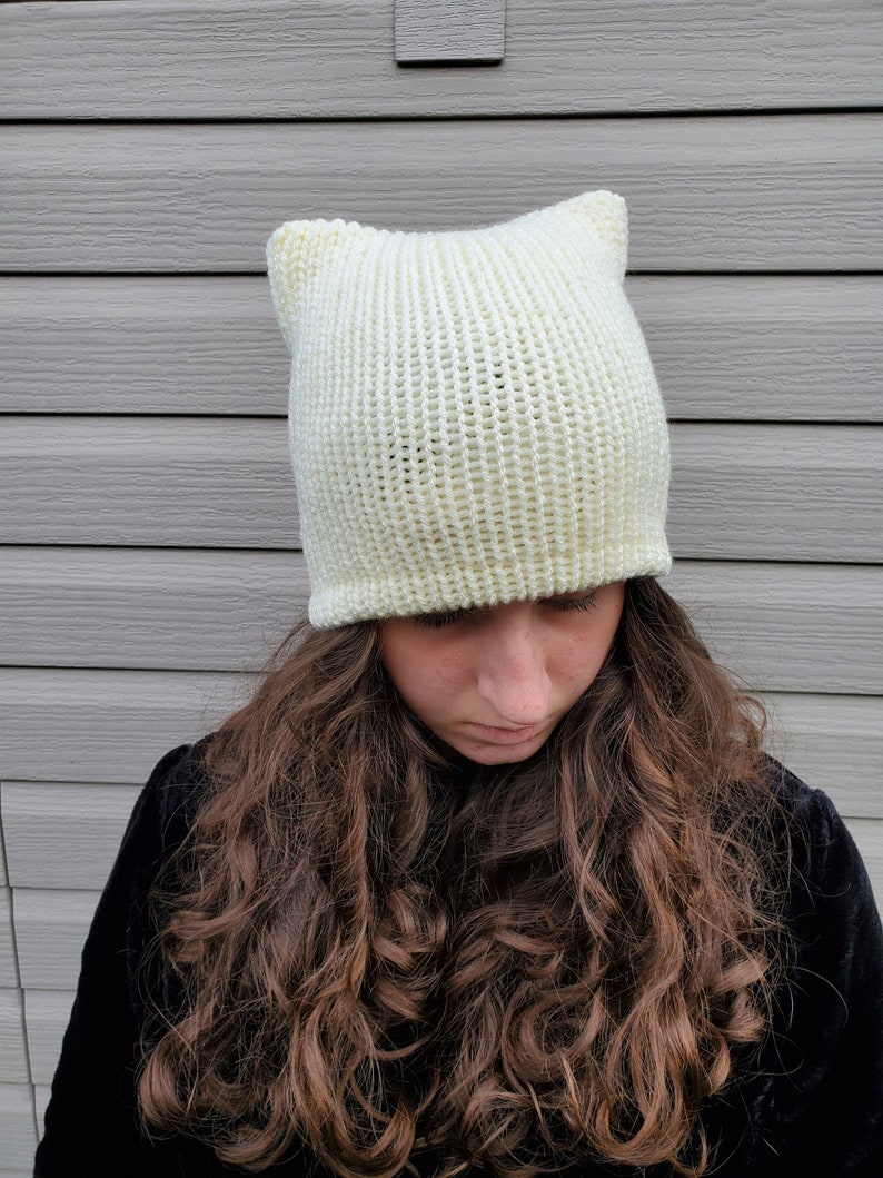 May include: A white knitted beanie with cat ears. The beanie is made of a soft, fluffy yarn and has a ribbed texture.