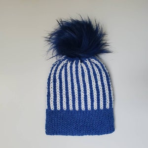 May include: A blue and white striped knit hat with a fluffy navy blue pom-pom on top. The hat features a solid blue brim and vertical white stripes on the main body. This winter hat is a cozy accessory.