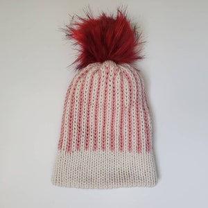 May include: A knitted winter hat with vertical stripes in cream and pink, topped with a fluffy, vibrant red pom-pom. The hat is a cozy accessory, perfect for cold weather. The pom-pom adds a playful touch to the design.