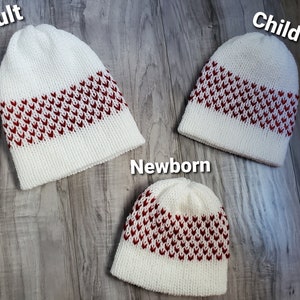 K&ouml;nnte beinhalten: Drei wei&szlig;e Strickm&uuml;tzen mit einem rot-wei&szlig;en Chevron-Muster. Die M&uuml;tzen sind mit "Erwachsene", "Kind" und "Neugeborenes" beschriftet.