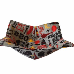May include: A grey fabric bowl cosy with a BBQ theme. The fabric is patterned with red, yellow, and black BBQ-related words and images, including a fork, sausage, and tomato. The bowl cosy is designed to hold a bowl.
