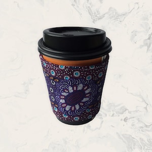 May include: A reusable coffee cup featuring a black plastic lid and a fabric sleeve. The sleeve is decorated with a purple, blue, and white dot pattern arranged in a circular mandala-like design, covering the middle section of the cup.
