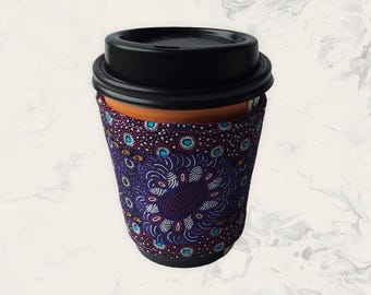 May include: A reusable coffee cup featuring a black plastic lid and a fabric sleeve. The sleeve is decorated with a purple, blue, and white dot pattern arranged in a circular mandala-like design, covering the middle section of the cup.
