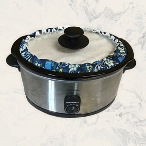 May include: A stainless steel slow cooker with a black rim and lid. A blue floral patterned cover with white trim is around the lid. The slow cooker has a black knob and the brand name "Sunbeam" is visible.