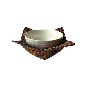 May include: A white ceramic bowl with horizontal ridges rests in a patterned bowl cosy. The cosy has a design of small, multicoloured circles on a dark background, with folded corners to hold the bowl.