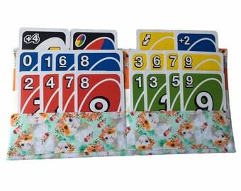 May include: A fabric card holder featuring a light green background with a repeating pattern of guinea pigs and small orange flowers. The holder contains multiple colorful numbered and action playing cards arranged in two rows within the pockets.