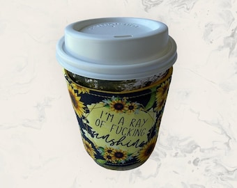May include: A reusable coffee cup sleeve featuring a sunflower pattern on a black background. The centre displays a yellow geometric shape with the text I'M A RAY OF FUCKING sunshine in black lettering. The sleeve is topped with a white plastic lid.