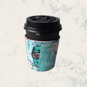 May include: A black disposable coffee cup with a black lid and a light blue fabric sleeve. The sleeve features a floral pattern and a graphic of a person wearing a surgical mask and goggles, with the text IN A WORLD FULL OF PRINCESSES BE A NURSE.