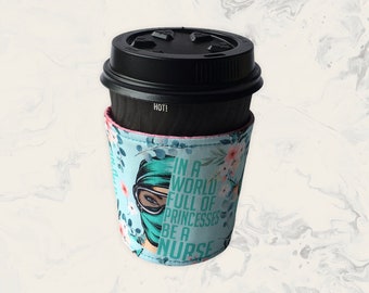 May include: A black disposable coffee cup with a black lid and a light blue fabric sleeve. The sleeve features a floral pattern and a graphic of a person wearing a surgical mask and goggles, with the text IN A WORLD FULL OF PRINCESSES BE A NURSE.