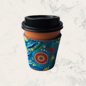 May include: A reusable coffee cup featuring a solid terracotta-coloured body and a black plastic lid. The cup is wrapped in a fabric sleeve decorated with a vibrant, multi-coloured pattern of concentric circles, leaves, and dots in shades of blue.