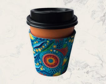 May include: A reusable coffee cup featuring a solid terracotta-coloured body and a black plastic lid. The cup is wrapped in a fabric sleeve decorated with a vibrant, multi-coloured pattern of concentric circles, leaves, and dots in shades of blue.