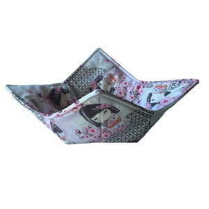 May include: A square fabric bowl cosy featuring a repeating pattern of stylised figures with black hair and pink cherry blossoms on a light background, accented by sections of a black and white geometric grid print with stitched edges.