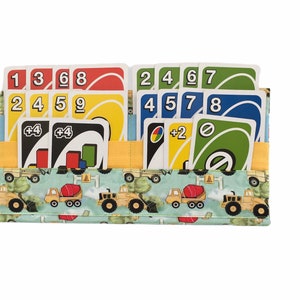 Peut inclure: Un porte-cartes en tissu sur le thème de la construction. Le porte-cartes a des poches pour les cartes et un rabat qui se ferme avec une fermeture velcro. Le porte-cartes est décoré d'un fond bleu et vert et de véhicules de chantier jaunes. Le porte-cartes est rempli de cartes UNO.