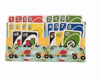 May include: A fabric card holder with a construction theme. The holder has pockets for cards and a flap that closes with a velcro strap. The holder is decorated with a blue and green background and yellow construction vehicles. The holder is filled with UNO cards.