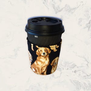Peut inclure: Un manchon de tasse à café noir orné d'un motif répétitif de chiens dorés sur fond noir. Le manchon entoure une tasse à café jetable noire avec un couvercle en plastique noir sur lequel est imprimé le mot HOT! sur le côté.