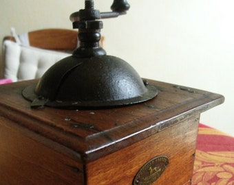 May include: A vintage wooden coffee grinder with a dark metal top and handle. The wooden box is a warm brown color, and a small metal plaque is affixed to the side. The grinder sits on a red patterned tablecloth.