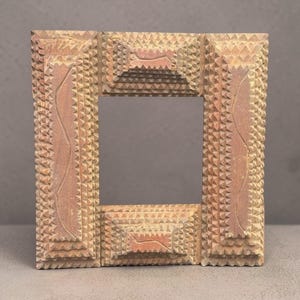 May include: Square wooden frame with a small mirror. The frame is a light brown color with a textured, geometric design. The corners and sides have a stepped appearance, and the frame features carved details.