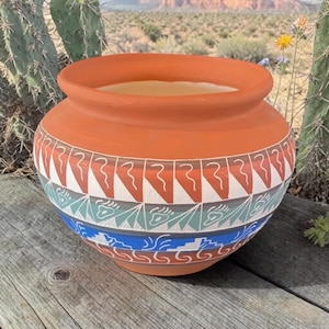 May include: A terracotta-colored ceramic pot with a wide opening and a rounded body. The pot is adorned with geometric patterns in white, red, blue, and green. The design includes triangles, wavy lines, and stylized shapes, creating a Southwestern aesthetic.