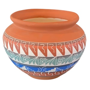 May include: A terracotta-colored ceramic pot with a wide opening and rounded body. The pot features a band of geometric patterns in white, red, blue, and teal. The design includes triangles, wavy lines, and stylized shapes. The pot is likely handmade.